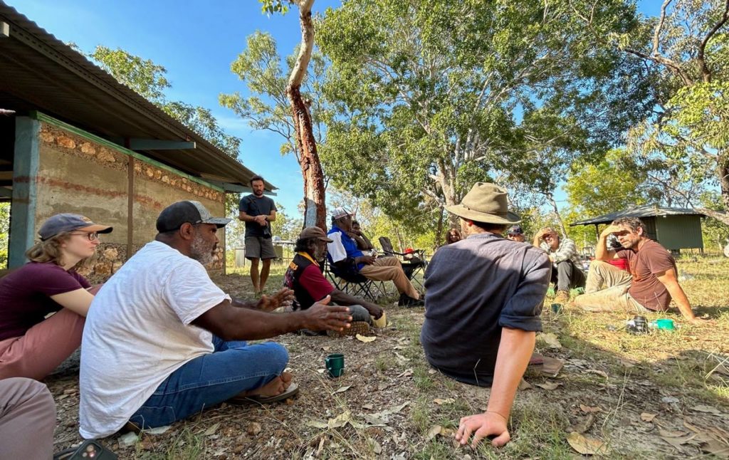 Jimmy Frank, seated, speaking and gesturing to a group of people sitting and standing in a rough circle next to a timber-framed, stone-walled house at Barapunta