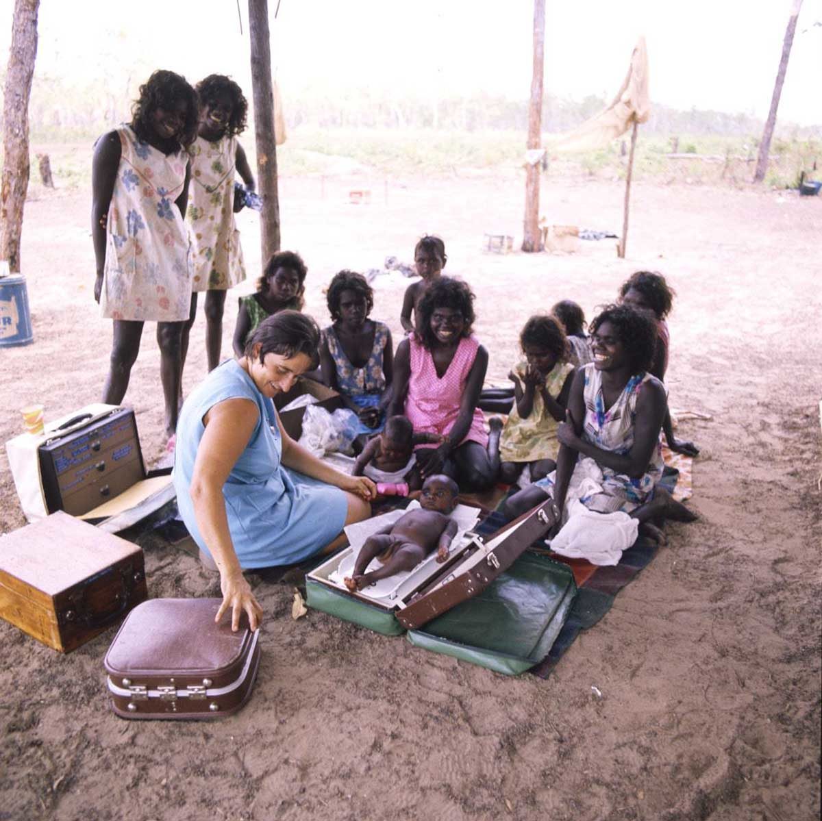 Gan-Gan-nurse-weighing-baby_A8598-AK5-3-80-42_1200 A balanda woman kneels on a sandy ground weighing an Aboriginal baby. Five Aboriginal women and four of their children are seated in front. Another two women are standing looking on.