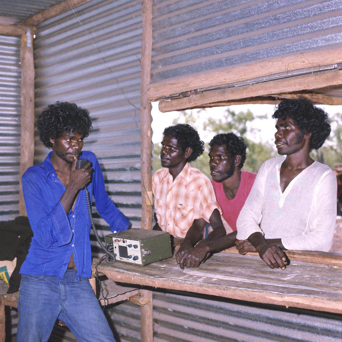 Gurrumurru_radio_A8598-AK29-4-80-14_1200 A young Aboriginal man stands talking into a radio in a bush-pole and corrugated iron structure with an open window. Standing outside at the window are three more young Aboriginal men, looking at the man inside.