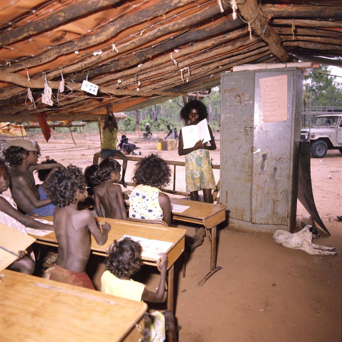 Homeland-school_A8598-AK5-3-80-14_1200 Under a hand-built open-sided timber shelter, a young Aboriginal woman stands showing an open book to a class of at least eight young students who are sitting and standing behind small desks. Next to the teacher is a tall metal cabinet. A dog lies in front of the cabinet and in the background is a land rover and several seated adults.