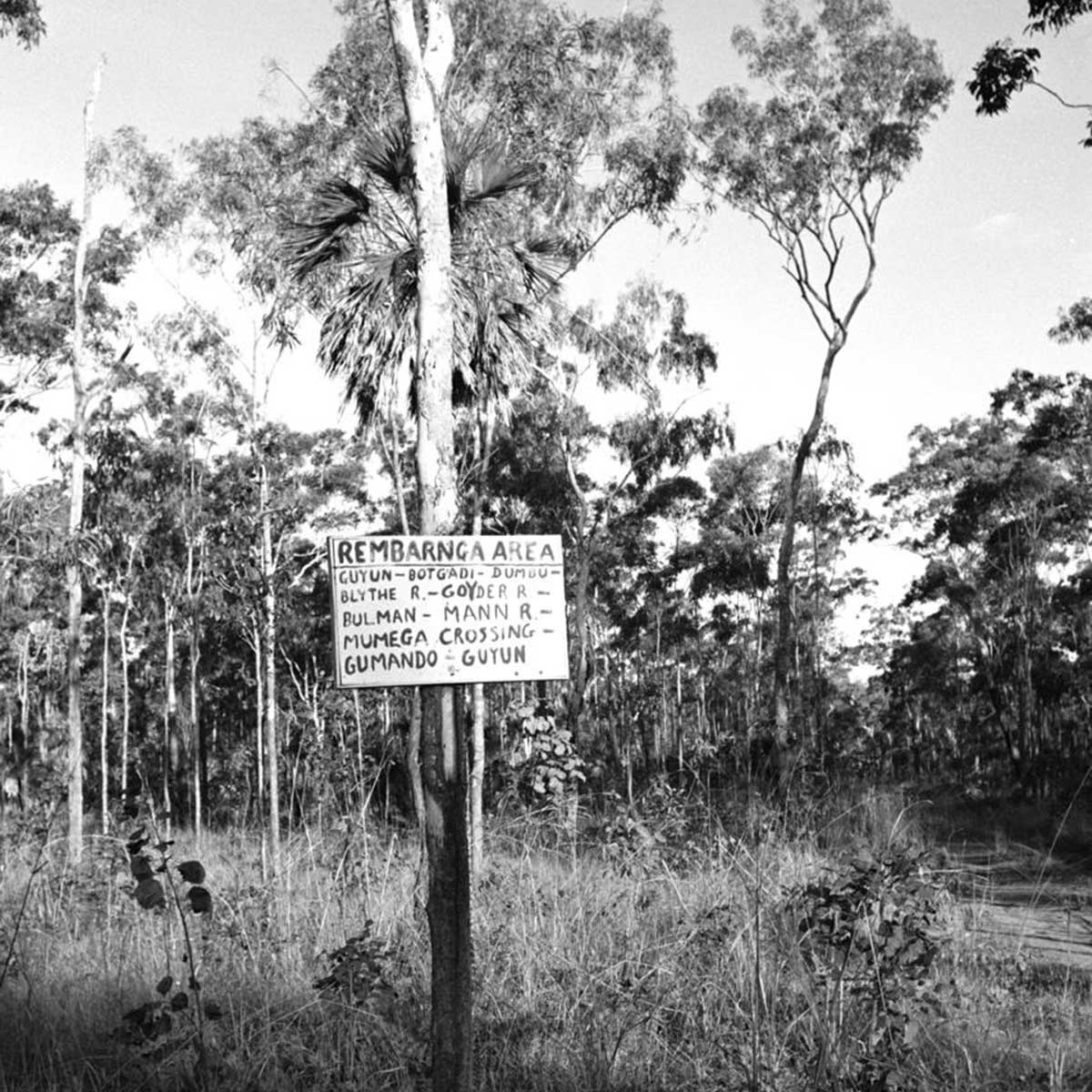 NAA_ItemNumber11645600_Rembarnga-area_1200 Bush sceen with a hand-painted sign fixed to a tree declaring it 'REMBARNGA AREA' and naming places within: Guyun, Botgaci, Dumou, Blythe R, Goyder R, Bulman, Mann R, Mumega Crossing, Gumando, Guyun