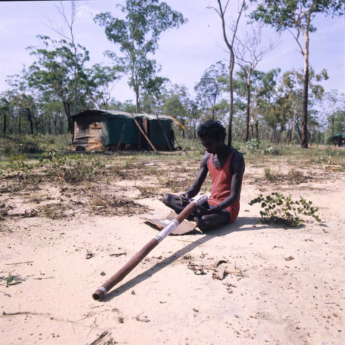 Painting-yidaki-outside-house_Mangallod_A8598-AK5-3-80-10_1200 A young Aboriginal man sits cross-legged on sandy ground painting a yidaki using white ochre on a bark plate. Behind him is a bark shelter and bushland.
