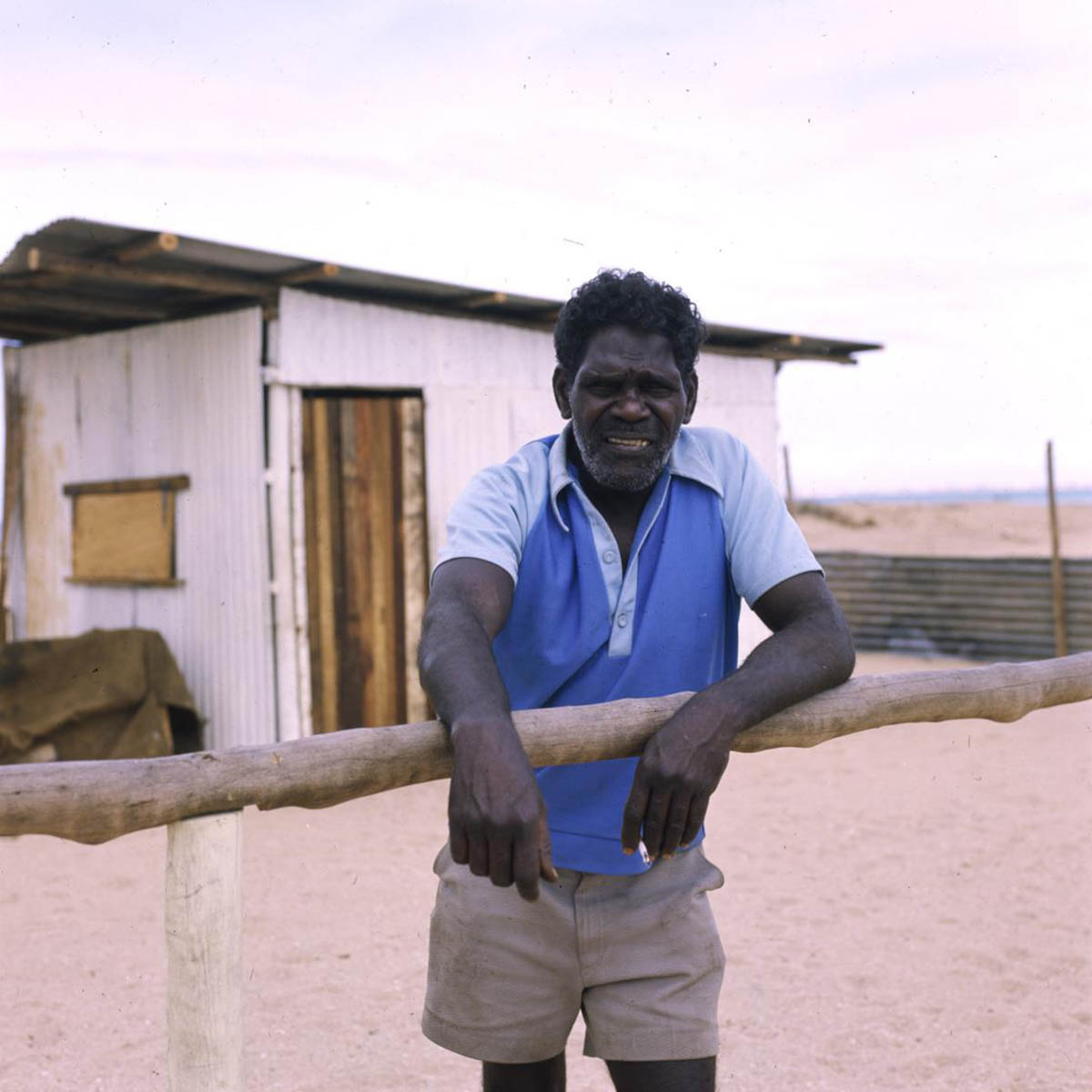 Yanbagwa_clan-leader_A8598-AK27-3-80-26_1200 Senior Aboriginal man standing on sandy ground, facing the camera, arms leaning on a bush-pole rail. Behind him is a hand-built one-room house.